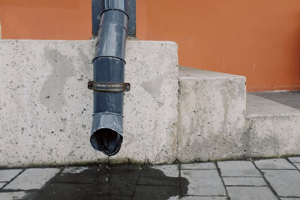 Metal drain pipe with water drops located on red wall near concrete stairs of modern building on street in city